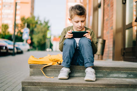 Back to school. Cute child with backpack, holding mobile phone, playing with cellphone. School boy pupil with bag. Elementary school student after classes. Kid sitting on stairs outdoors in the streetの写真素材
