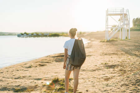 Young Female volunteer with bag and gloves picking up trash, a plastic bottles, clean up beach with a sea. Woman collecting garbage. Environmental ecology pollution concept. Earth Day.の写真素材
