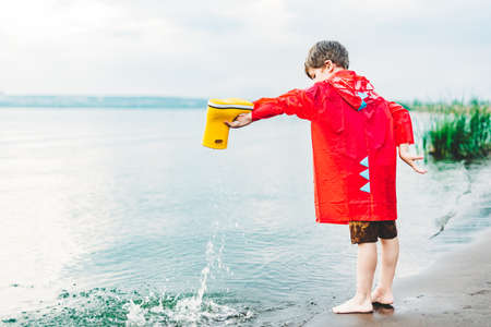 Boy in a red raincoat pours water out of yellow rubber boot into the lake. Child playing with water at pond. Kid having fun outdoors near river bank.の写真素材