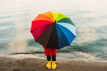 Boy in a red raincoat and yellow rubber boots holds rainbow umbrella standing at beach. Child with colourful umbrella looking at sea. School kid in a waterproof red coat holding multicolor umbrella.の写真素材