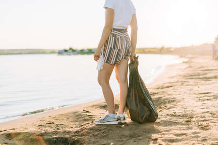 Cropped Female volunteer with bag and gloves picking up trash, a plastic bottles, clean up beach with a sea. Woman collecting garbage. Environmental ecology pollution concept. Earth Day.の写真素材