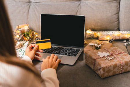 Close up of woman hands with credit card, gifts, and mock up screen laptop. Online shopping at Christmas holidays. Cropped female sit on couch with natural eco presents and decor. Merry Christmas.の写真素材