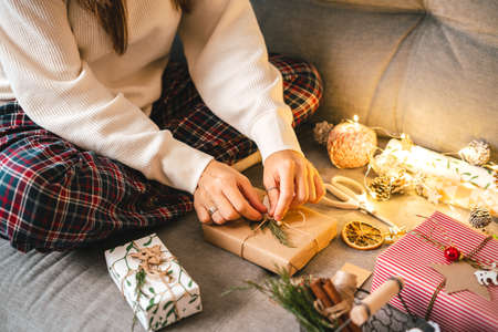 Woman s hands wrapping Christmas gift boxes, close up. Cropped female sit and preparing natural eco presents on couch with decor elements and items. Merry Christmas or New year DIY packing Concept.の写真素材