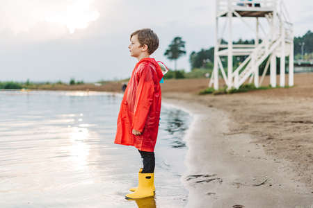 Boy in a red raincoat and yellow rubber boots playing with water at the beach. School kid in a waterproof coat jumping in water at sea. Child having fun with waves at the shore.の写真素材