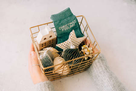 Golden basket in woman hands with eco decor for Christmas gift. Space for text. Preparing care package, seasonal gift box with socks, wooden house, paper toys. Zero waste, plastic free.の写真素材