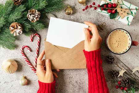 Desktop of woman hands in red sweater holding blank paper and envelope . Flat lay of gray background with cup of coffee and Christmas decoration. Top view mock up and copy space for text.の写真素材