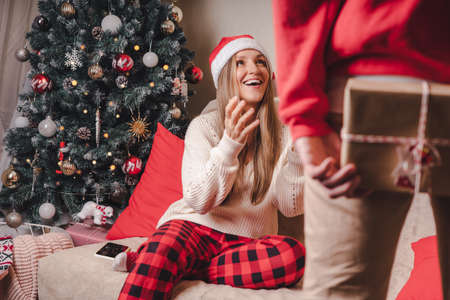 Surprising her... Close-up rear back view of man in red sweater holding a Christmas gift box behind his back while standing near decorated christmas tree. Family couple at Christmas holidaysの写真素材