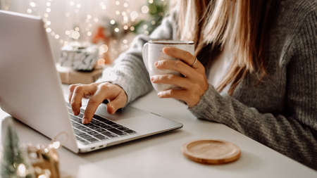 Close up of woman hands with, gifts, coffee cup and laptop. Online shopping at Christmas holidays. Freelance girl woking from home office. Female typing at notebook computer. Christmas moments.の写真素材