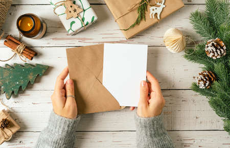Woman hands in sweater holding blank paper and envelope . Flat lay of white wooden background with...