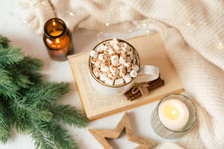Hygge Cup of hot cocoa or coffee with marshmallow, sweater, candle, book, cinnamon and fir tree. Christmas lights cozy background. Xmas holiday wooden star decorations.の写真素材