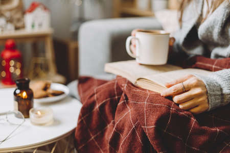 Cozy woman legs in knitted winter warm sweater and checkered plaid drinking hot cocoa or coffee in mug, reading book, during resting on couch at home. Christmas holidays with decor and lightsの写真素材