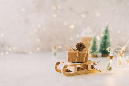 Eco Natural Christmas gift on toy wooden sled on white background with glowing light bokeh, copy space. New year card. Xmas decoration with house, Christmas tree and snowの写真素材