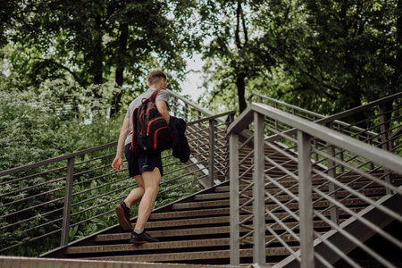 Tired Young man runner in headphones and backpack walk and relaxing after sport training. Holding water bottle while doing fitness workout in summer sunny green park.の写真素材