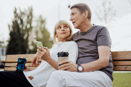 An adult mature happy couple in love sitting on bench with phone outdoors in park. A blonde caucasian man and woman spend time together and drinking coffee. Senior wife and husband walking outsideの写真素材