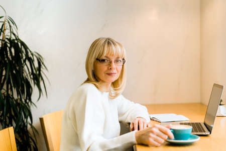Mature adult woman sitting in cafe with coffee mug and working online on laptop. Businesswoman in eyeglasses typing on notebook computer in coworking space in roasters coffee shop.の写真素材