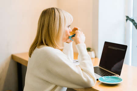 Mature adult woman sitting in cafe, drinking coffee mug and working online on laptop. Businesswoman in eyeglasses typing on notebook computer, in coworking space in roasters coffee shop.の写真素材