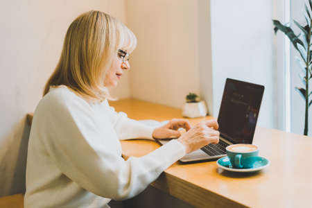 Mature adult woman sitting in cafe with coffee mug and working online on laptop. Businesswoman in eyeglasses typing on notebook computer in coworking space in roasters coffee shop.の写真素材