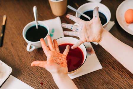 Easter day. Cropped close up Kid boy with dirty hands painting eggs on wooden background. Child sitting in a kitchen. Preparing for Easter, creative homemade decoration.の写真素材