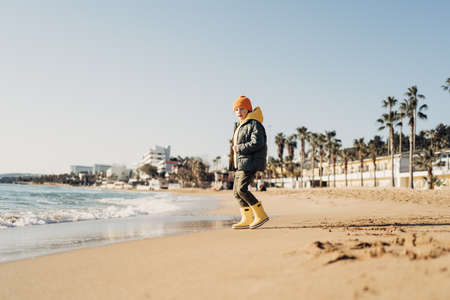 Boy in yellow rubber boots playing with water and sand at the beach. School kid touching water at autumn winter sea. Child having fun with waves at the shore. Spring Holiday vacation concept.の写真素材