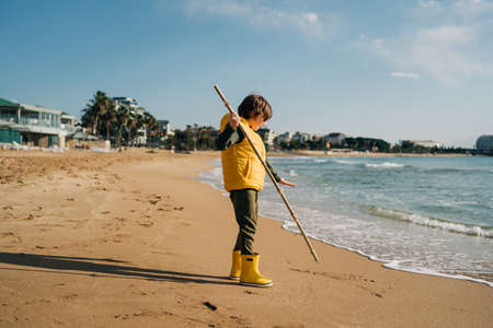 Boy in yellow rubber boots playing with stick and sand at the beach. School kid touching water at autumn winter sea. Child having fun with waves at the shore. Spring Holiday vacation concept.の写真素材