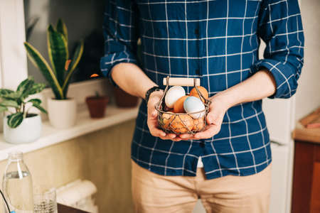 Easter day. Adult man holding basket with eggs on wooden background. Stay in a kitchen with nest. Preparing for Easter, creative homemade decoration.の写真素材