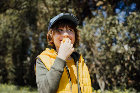 Smiling cheerful child kid in yellow vest and green hoodie eats crisp snacks outdoors in public...