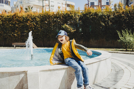 Schoolboy sitting on a fountain in public park during spring sunny day playing with water. Kid in yellow vest chilling in park with water sprinkling in the background. Summer holidays outside.の写真素材