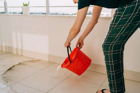 Man spilling the water from red bucket onto the tiled floor on the balcony in sunny day. Adult male doing home chores washing floors on the patio. Plant in a flower pot standing on balcony railings.の写真素材