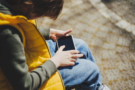 Schoolboy plays game his cell phone sitting on bench in the city park. Boy kid in yellow vest and green hoodie uses mobile gadget to chat with friends while outdoors in the garden during sunny day.の写真素材