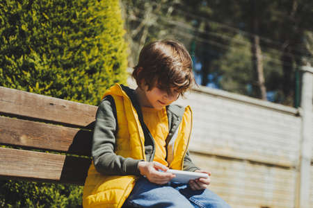 Schoolboy plays game his cell phone sitting on bench in the city park. Boy kid in yellow vest and green hoodie uses mobile gadget to chat with friends while outdoors in the garden during sunny day.の写真素材