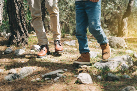 Close-up view of tourists school boy and his dad feet boots walking a stone footpath in spring forest. Child boy and father wearing hiking boots while walking in summer greenwood forest.の写真素材