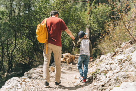 Rear view of tourists school boy and his dad walking a stone footpath in spring forest. Child kid and father wearing casual clothes and yellow backpack while hiking in summer greenwood leaf forest.の写真素材