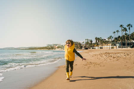 Boy in yellow rubber boots playing with sand at the beach. School kid touching water at autumn...