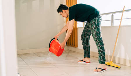 Man spilling the water from red bucket onto the tiled floor on the balcony in sunny day. Adult male doing home chores washing the floors on the patio. Broom standing against the balcony wall.の写真素材