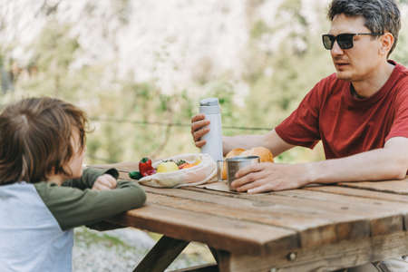 Close-up view of father and his school boy son on a family picnic in the mountains. Child kid and his dad taking a rest and enjoying a picnic while hiking in the mountains.の写真素材