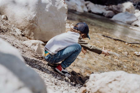 Caucasian school boy throwing rocks into the canyon river. Kid child playing with stones while sitting on a boulder near mountain river bank.の写真素材