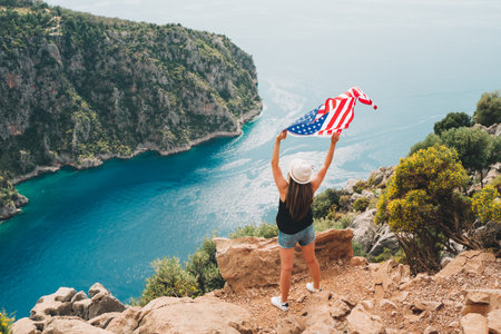 Young woman standing on a rock cliff and waving the US flag while looking at sea beneath. Girl traveller waving the American flag while standing on a mountain top. 4 fourth July Independence Dayの写真素材