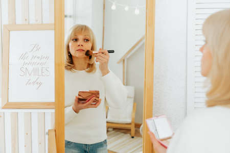 Beautiful smiling mature senior caucasian woman with blonde hair standing near mirror with make up palette and brush, doing makeup and looking at reflection in living room at home.の写真素材