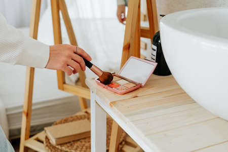 Close up of cropped caucasian woman with long hair holding powder palette and brush, doing makeup and looking at reflection in mirror at home.の写真素材