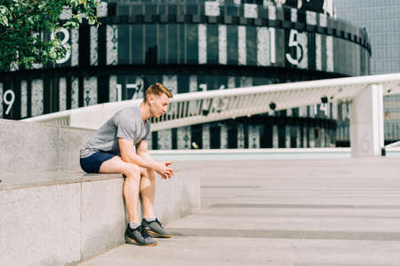 Tired Young man runner sitting on stairs and relaxing after sport training. Holding water bottle while doing workout in summer city street, cloudy skyの写真素材