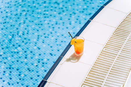 Tropical sparkling cocktail by the pool. The picture of glass with orange lemonade fruit cocktail standing near the poolside. Summer alcohol free drink by the hotel pool. Hello summer holiday vacationの写真素材