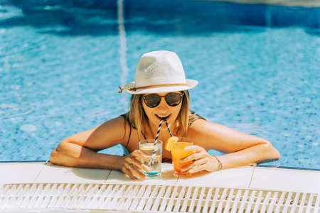 Portrait of beautiful cute smiling young woman lady girl in a bikini, hat, and sunglasses holding tropical lemonade cocktail while chilling in swimming pool. Hello summer holiday vacationの写真素材