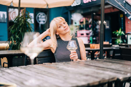 Mature adult woman sitting in bar outdoors with wine glasses and blurry restaurant background scene, drinking white wine and eating cheese. Summer sunny day on patio. People lifestyleの写真素材
