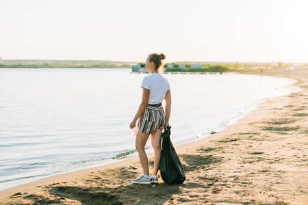 Young Female volunteer with bag and gloves picking up trash, a plastic bottles, clean up beach with a sea. Woman collecting garbage. Environmental ecology pollution concept. Earth Day.の写真素材