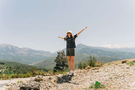 Young woman in hat standing on Mountain View. Raised hands on mountain background.の写真素材