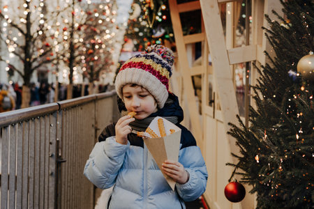 Cute little boy eating churros on traditional Christmas fair in snow. Child kid enjoying sweets on Xmas market. Winter holidays with family. Merry Christmas.の写真素材