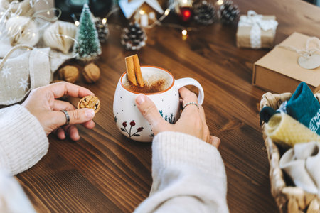 Christmas mug of coffee with cinnamon in woman hands. Wrapping Christmas eco gift box, close up. Preparing presents on wooden table with natural decor elements and items Christmas packing Concept.の写真素材