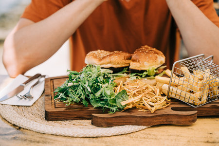 Cropped closeup man male sitting in front of sliced grilled tasty beefy cheese burger hamburger and french fries on a plate in the street cafe. Copy blank space.の写真素材