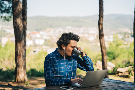 Happy cheerful man with a laptop sitting outdoors in nature forest park. Freelancer with computer typing, blogging, browsing in vacation in mountains. Freelance, distance work concept.の写真素材