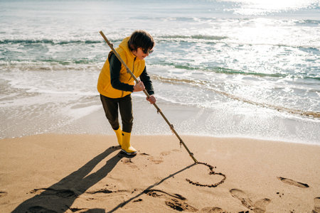 Boy in yellow rubber boots drawing heart shape on sand at the beach. School kid touching water at autumn winter sea. Child having fun with waves at the shore. Spring Holiday vacation concept.の写真素材